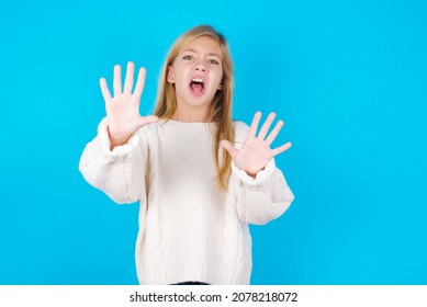 Dissatisfied Caucasian Little Kid Girl Wearing White Knitted Sweater Over Blue Background Frowns Face, Has Disgusting Expression, Shows Tongue, Expresses Non Compliance, Irritated With Somebody.