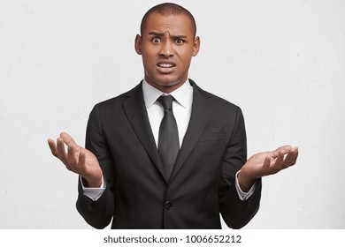Dissatisfied dark skinned male manager with puzzled expression, clenches teeth, being hesitant and frustarted, recives notice to terminate contract, dressed in formal suit, poses against white wall