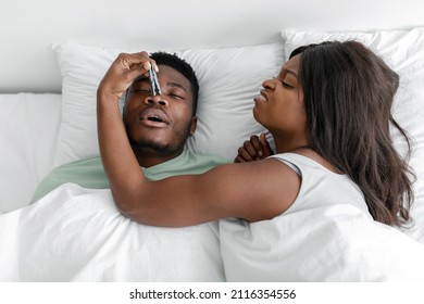 Dissatisfied funny millennial black lady wake up from snoring sleeping man, closes his nose with clothespin, lies on white bed in bedroom, top view. Noise, sleep problems, insomnia, snoring remedy