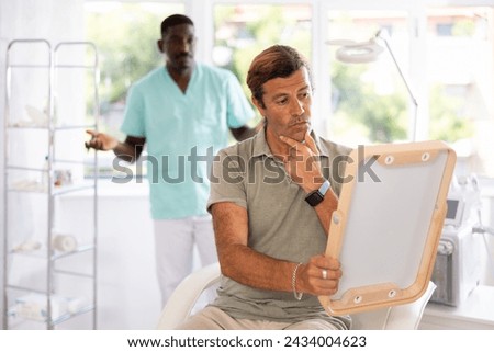 Dissatisfied man, sitting on examination chair and skeptically scrutinizing face in mirror after cosmetic procedures while bewildered cosmetologist making confused hands gesture in blurry background