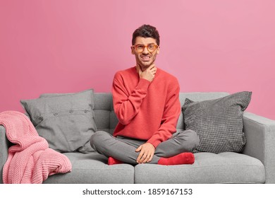 Dissatisfied man touches sore throat has inflammation sits crossed legs on cozy sofa stays at home as being ill wears casual clothing isolated on pink background. Unhealthy guy has symptoms of cold