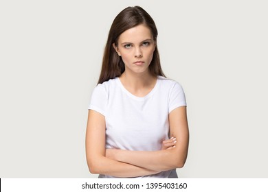 Dissatisfied millennial brown haired woman arm crossed looks at camera posing on grey background, disgruntled girl wearing white t-shirt irritated face expressions show negative attitude concept image