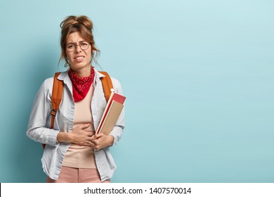 Dissatisfied Schoolgirl Has Stomachache After Eating Spoiled Food In School Canteen, Smirks Face In Displeasure, Dressed In Casual Clothes, Holds Notebooks Isolated On Blue Background. Bad Feeling