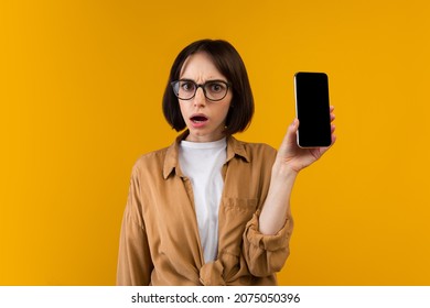 Dissatisfied Shocked Lady Holding Smartphone With Blank Screen, Showing App Or Website, Being Disappointed, Standing Over Yellow Background, Studio Shot, Mockup