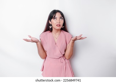Dissatisfied young Asian woman dressed in pink blouse rolling her eyes and raising hand discontentedly, standing against white background
