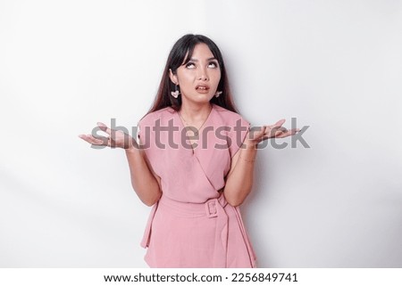 Dissatisfied young Asian woman dressed in pink blouse rolling her eyes and raising hand discontentedly, standing against white background