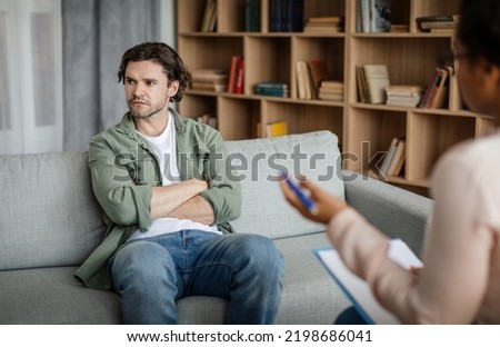 Dissatisfied young caucasian guy client ignores doctor at meeting with black lady psychologist in office clinic interior. Denial and psychological therapy, session with professional, help and support