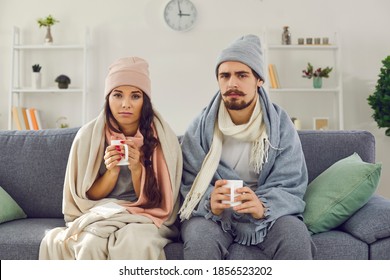 Dissatisfied young couple having problem with central heating, sitting on sofa at home, freezing, drinking hot tea trying to warm up. Sick man and woman wrapped in blankets suffering from cold or flu - Shutterstock ID 1856523202