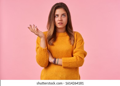 Dissatisfied young pretty brown haired woman dressed in mustard knitted sweater rolling her eyes and raising hand discontentedly, standing against pink background