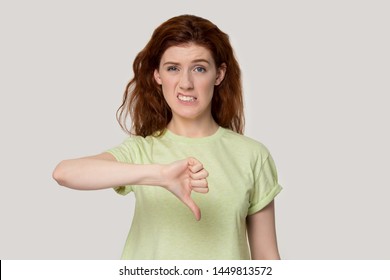 Dissatisfied young red-haired woman showing thumbs down gesture, unhappy female customer giving negative feedback on product or service, does not recommend store, isolated on studio background.