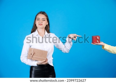 Dissatisfied young woman in elegant outfit, loathingly pointing at gift. Crop view of anonymous hand in shirt holding small wrapper present box, isolated on blue studio background. Concept of emotion.