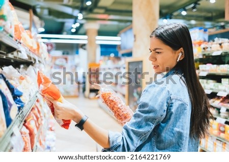 Dissatisfied young woman wearing casual outfit and earphones holding two packs of pasta looking at label while standing in grocery department in supermarket