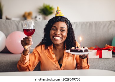 Distance Birthday Party. Cheerful African American Lady Holding B-Day Cake With Candle And Glass Of Wine Sitting At Home. Woman Posing Wearing Festive Hat Celebrating Holiday Online Via Video Call