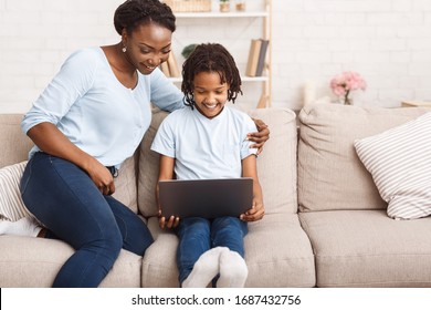 Distance Education Concept. African mother helping her child with homework, using laptop, sitting on sofa at home. Free space