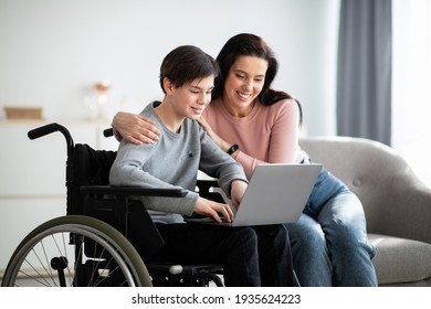 Distance Education. Joyful Disabled Teenager In Wheelchair Using Laptop, Making Homework With His Mother Indoors