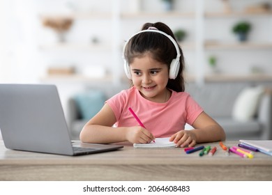 Distance Education. Little Arab Female Child Wearing Wireless Headphones Study With Laptop At Home, Smiling Cute Girl Drawing With Pencils In Notepad And Looking At Computer Screen, Free Space - Powered by Shutterstock