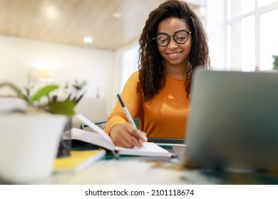Distance Education. Portrait of smiling black lady in glasses sitting at desk, using laptop and writing in notebook, taking notes, watching tutorial, lecture or webinar, studying online at home - Shutterstock ID 2101118674