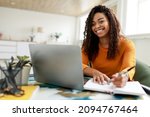 Distance Education. Portrait of smiling woman sitting at desk, using laptop and writing in notebook, taking notes, watching tutorial, lecture or webinar, studying online at home looking at screen