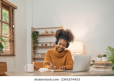 Distance Education. Portrait of smiling woman african american sitting at desk, using laptop and writing in notebook, taking notes, watching tutorial, lecture or webinar, studying online at home - Powered by Shutterstock