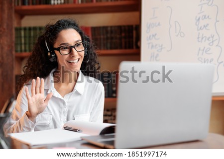 Distance Education. Positive lady wearing glasses and wireless headest at virtual meeting, sitting at desk, having video call on laptop, waving to webcam. Woman studying or teaching online at home