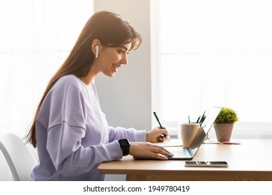 Distance Education. Profile of smiling woman in wireless headphones sitting at desk, using laptop and writing in notebook, taking notes, watching tutorial, lecture or webinar, studying online at home