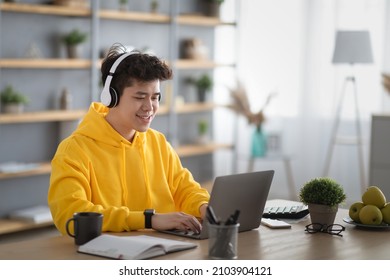 Distance Education. Smiling Asian Man In Wireless Headset Using Laptop At Home, Typing On Pc Keyboard, Cheerful Male Student Having Online Lesson Watching Webinar Or Doing Homework Sitting At Desk