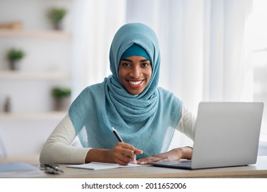 Distance Job. Portrait Of Happy Black Muslim Lady In Hijab Taking Notes While Working With Laptop At Home Office, Smiling African Islamic Female Sitting At Desk With Computer And Looking At Camera