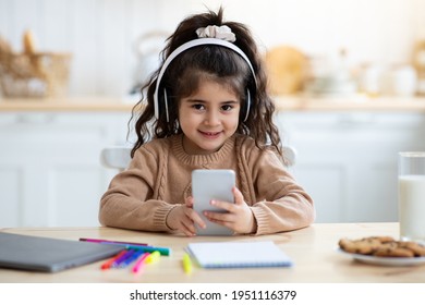 Distance Learning. Adorable Arab Girl Study With Smartphone And Wireless Headphones In Kitchen, Having Online Lesson Or Playing Mobile Games While Sitting At Table At Home, Closeup With Free Space