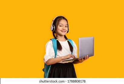 Distance Learning. Asian School Girl With Laptop And Headphones Listening To Educational Podcast Studying Standing Over Yellow Studio Background.