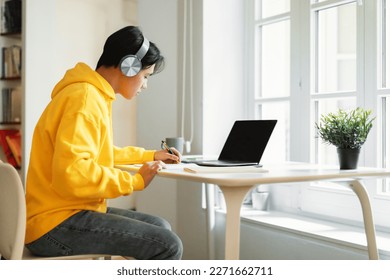 Distance Learning. Asian Teenager Boy At Laptop Computer Writing Taking Notes Doing Homework Online Sitting At Table At Home, Wearing Wireless Headphones. Side View Shot - Powered by Shutterstock