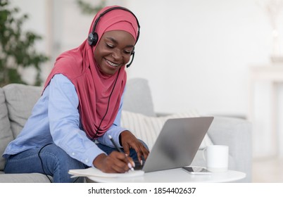 Distance Learning. Black Lady In Hijab Studying Online With Laptop At Home, Watching Webinar And Taking Notes, African Muslim Woman Wearing Headset, Sitting On Couch In Living Room, Copy Space - Powered by Shutterstock