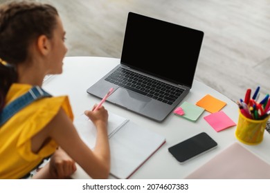 Distance Learning. Happy Girl Using Laptop With Black Blank Screen Mock Up At Home, Studying Online With Computer, Female Child Sitting At Desk And Writing In Notebook, Rear Back Over Shoulder View