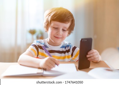 Distance Learning Online Education. Caucasian Smile Kid Boy Studying At Home With Smartphone And Doing School Homework. Thinking Child Siting At Table With Cell Phone Due Coronavirus Quarantine