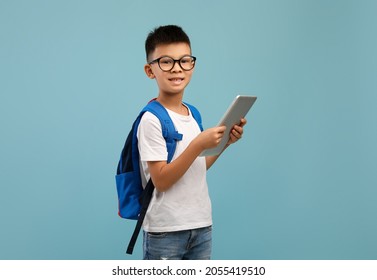 Distance Learning. Smiling Asian Schoolboy Wearing Eyeglasses And Backpack Holding Digital Tablet, Korean Male Child Using Modern Technologies For Studying Online, Standing On Blue Studio Background