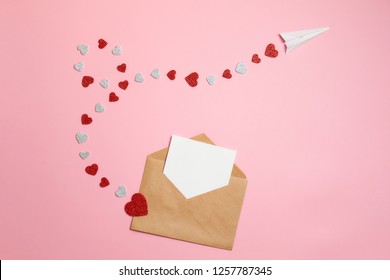 Distance Love Concept, Sending Love Letter, Valentines Day. Kraft Envelope With Blank Postcard And Paper Airplane Flying On Route Made Of Heart Shaped Valentines Cards Lay On Pink Background Desk