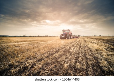 Distance shot of modern red tractor using navigation during seeding directly into the stubble after the harvest with dramatic sky during autumn day.