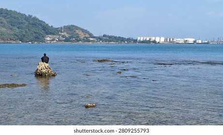 Distant back view of an unrecognizable person sitting on a rock in the calm, crystal-clear blue shallow ocean water near the beach during a bright sunny day.