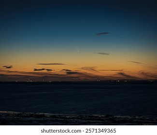 A distant comet in the evening sky at sunset over an estuary. The clouds are dramatic, and there are city lights on the horizon. The comet is visible, but subtle.