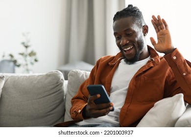 Distant Communication Concept. Excited Black Guy Making Video Call Via Cellphone, Sitting On Couch At Home And Talking With Friends Of Family, Waving Hand To Phone Webcam