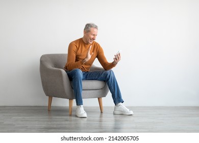 Distant Communication Concept. Portrait Of Happy Mature Male Making Having Video Call Sitting On Chair At Home And Talking With Friends Of Family, Waving Hand To Cell Phone Webcam, White Studio Wall