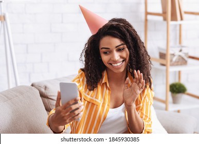 Distant Communication And Congratulation. Portrait Of African American Lady Holding Mobile Phone, Waving To Webcam, Wearing Party Hat, Celebrating Virtual Birthday At Home. Remote Meeting