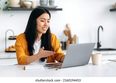Distant communication. Friendly pretty asian brunette girl, manager, freelancer, sit in the kitchen, talking on video call by laptop with the client or employees, discussing project, smiling - Shutterstock ID 2160194453