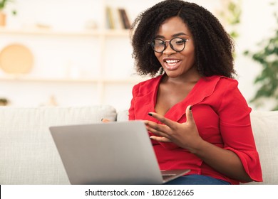 Distant Communication. Happy african woman in glasses and red shirt making video call sitting on couch and talking - Powered by Shutterstock