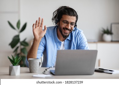 Distant Communication. Happy Western Man In Headset Waving Hand At Laptop Camera While Making Video Call Via Computer In Office, Chatting With Family Or Having Online Conference, Copy Space