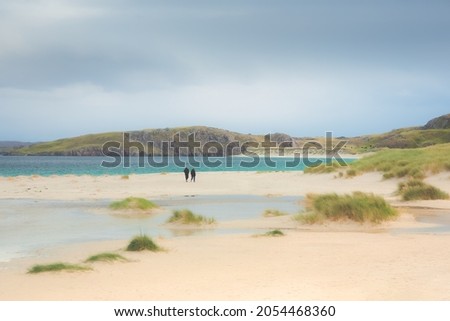 A distant couple take a leisurely stroll along the white sand beach and grassy dunes at Reef Beach on the Isle of Lewis and Harris in the Outer Hebrides of Scotland, UK.