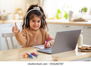 Distant Education. Adorable Little Girl In Headphones Using Laptop In Kitchen, Sitting At Table And Showing Thumb Up At Camera, Female Child Study Online With Computer And Drawing With Pencils - Powered by Shutterstock