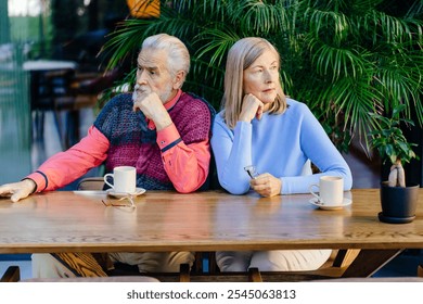 Distant elderly couple sitting at cafe table, cups served. Older man and woman sit side by side, both looking away. Concept of relationship stress, quarrel. Cafe surrounded by greenery. - Powered by Shutterstock