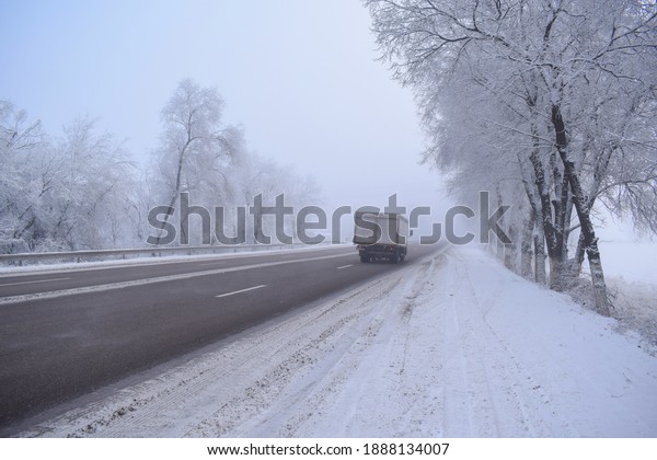 Distant\
lorry on a winter road with thick fog. Early morning winter highway\
with frost-covered trees on both sides of the road and a lonely car\
or lorry moving away. Car on winter highway\
road.