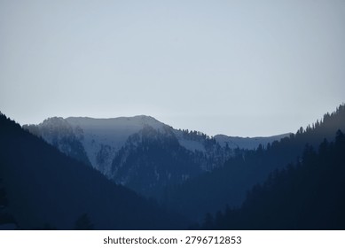 A distant mountain ridge rises in the background, its crest partially covered in pale patches of winter snow. The icy slopes are nestled between rugged rocky peaks that sit quietly along the horizon line. A soft, dim light illuminates the very top edge of the snowy ridge, while the deeper crevices and lower rock faces remain cast in cold blue shadow.

In the foreground, two steep mountain slopes covered in dense pine forests descend from the left and right sides to frame the deep valley below. These nearby forested ridges appear as dark, solid silhouettes against the faint background landscape. The sky above is entirely clear and displays a very light, pale blue gradient, creating a calm, muted atmosphere over the remote mountain wilderness.