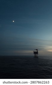 A distant offshore platform stands beneath a twilight sky with the moon visible overhead. The minimalist composition combines offshore industry with the peaceful atmosphere of dusk.